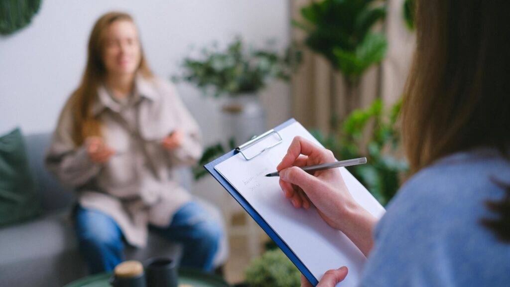 A close-up of a person holding a clipboard and pen, taking notes during a conversation. In the blurred background, another person is sitting on a couch, gesturing with their hands, surrounded by plants and a cozy setting.